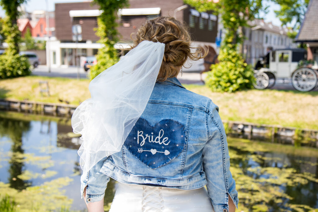 Bruid met sluier die wappert in de wind en een spijkerjasje waar "bride" op staat geborduurd